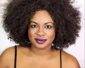 Headshot of Tanika Baptiste. She is a Black woman with shoulder length natural curly brown hair. They are staring directly at the camera and wearing purple lipstick.