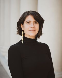 Headshot of Amal Bisharat. She is standing in front of white columns and wearing a black turtleneck shirt and one gold earring. She is a light skinned Palestinian American woman with chin length dark brown hair.