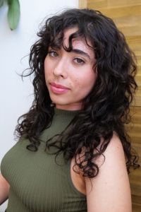 Headshot of Mylo Cardona. They are leaning against a yellow and white wall. They are wearing a green sleeveless shirt and looking toward the camera at an angle. They have curly medium length hair with bangs.
