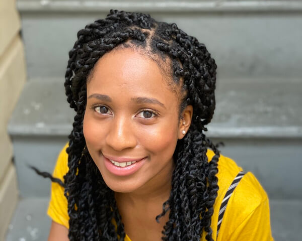 Angel sits smiling on a grey staircase wearing a yellow shirt and black and white striped pants. She is a black woman with waist length dark hair styled in twists.