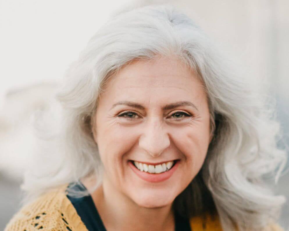 Headshot of Debórah Eliezer. She is a white woman with white hair wearing a black shirt with a yellow sweater. She is looking at the camera and smiling with her teeth showing.