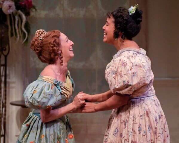 Two women in costume looking at each other holding hands and laughing. The woman on the left is a white woman with red hair in a bun with curls wearing a light blue dress with puffy sleeves. The woman on the right has light brown skin and dark brown hair in a curly bun. She is wearing a white dress with blue flowers and puffy sleeves.
