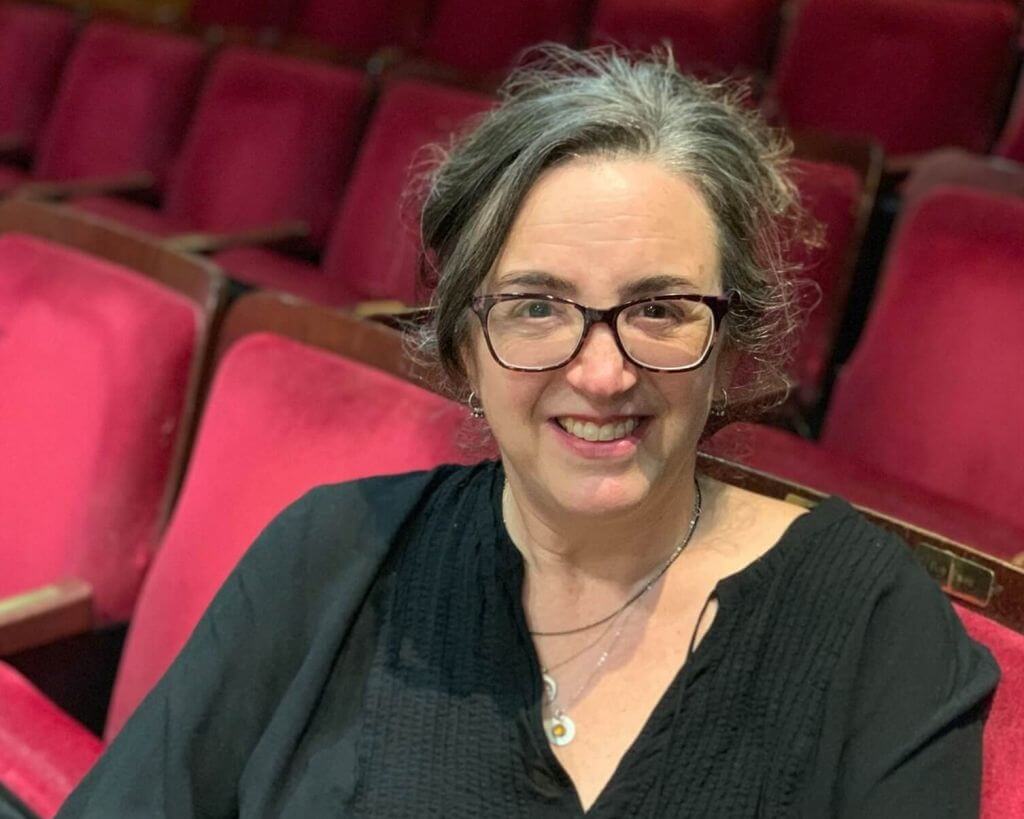 Cindy Goldfield sitting in red theatre seats. She is a light skinned woman with salt and pepper hair swept back from her face. She is wearing glasses and a black shirt and three silver necklaces.