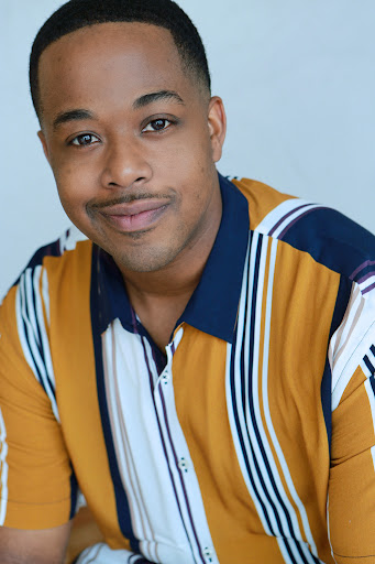 Headshot of Devin Cunningham. He is a light skinned black man with short black hair with a nice fade and clean edges and a small moustache. He is wearing a color block button up shirt that has thick and thin dark blue , mustard yellow and white stripes. 