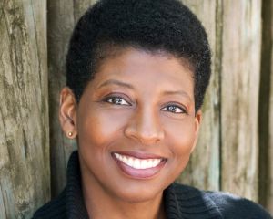 Headshot of Velina Brown. She is a black woman with short cropped hair. She is smiling with her teeth showing. She is wearing a black shirt and leaning against a wood paneled background.