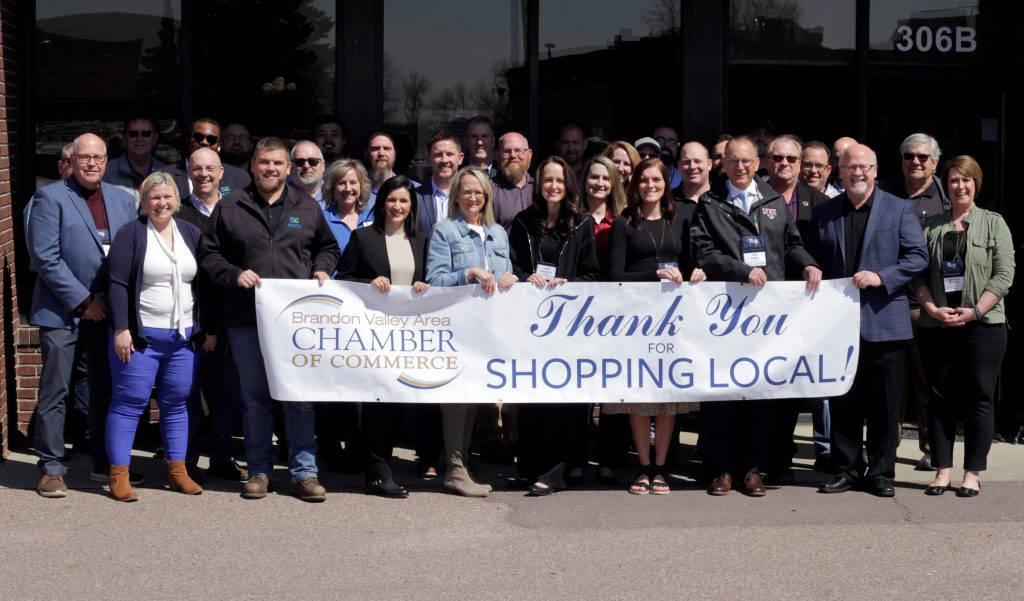 A large group of people pose under a blue awning that says "Brandon Valley Area Chamber of Commerce. They hold a banner that reads: "Thank you for Shopping Local!" and has the BVA Chamber Logo in blue and gold.