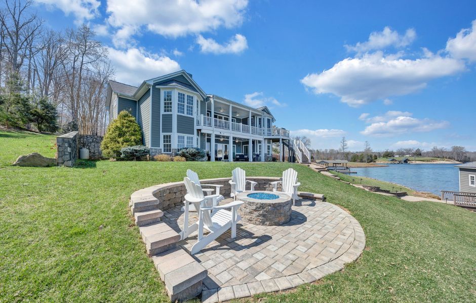 Four white chair surround an outdoor firepit with a large gray lakefront house in the background